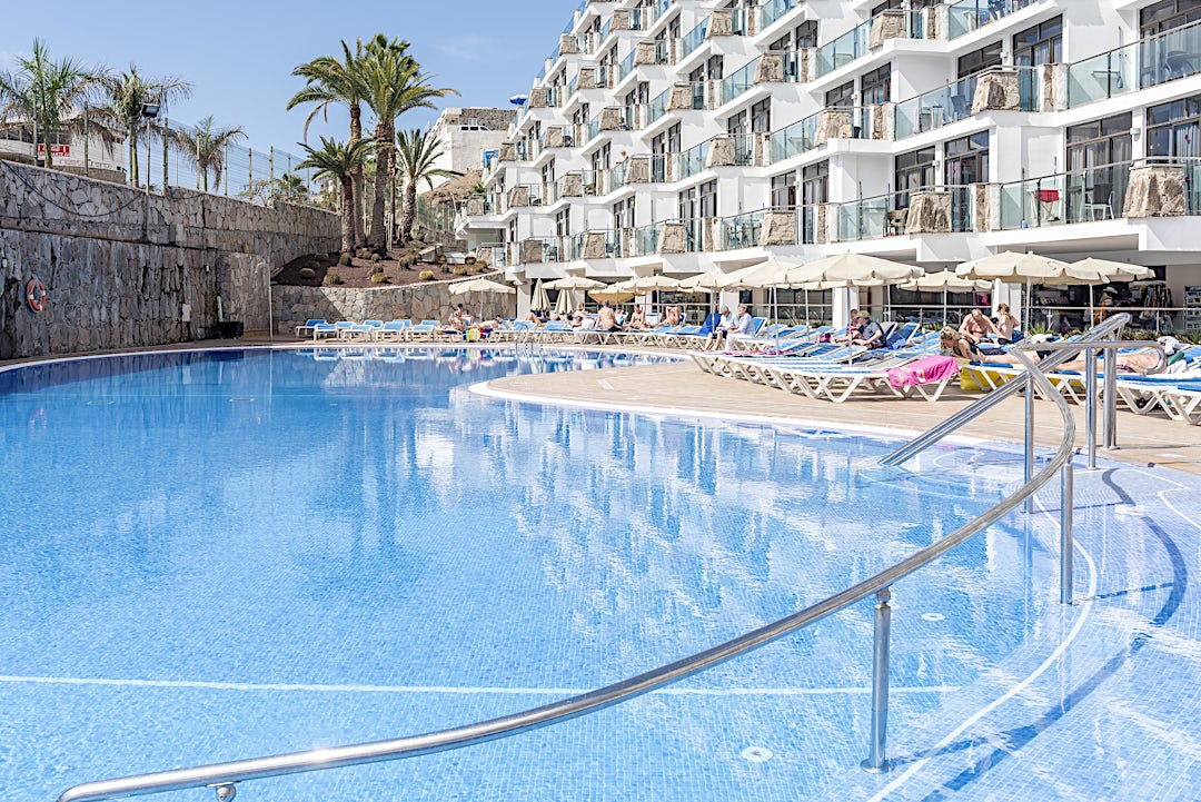 Bassengområdet med solsenger, parasoller og hotellets fasade på den ene siden og en steinmur på motsatt side.
