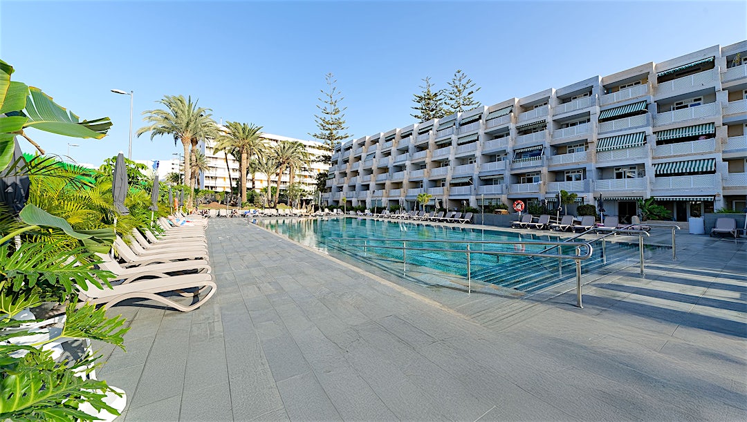 Hotellets fasade, basseng og solterrasse med solsenger og parasoller omringet av palmer.