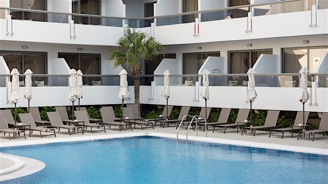 Bassengområde med solsenger og parasoller ved bassenget og hotellets hvite fasade med balkonger i bakgrunnen.