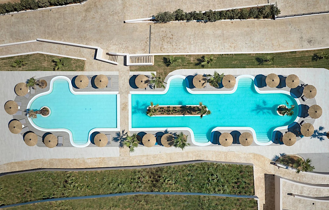 Fugleperspektiv av hotellet med turkist basseng, solsenger og grønne planter.